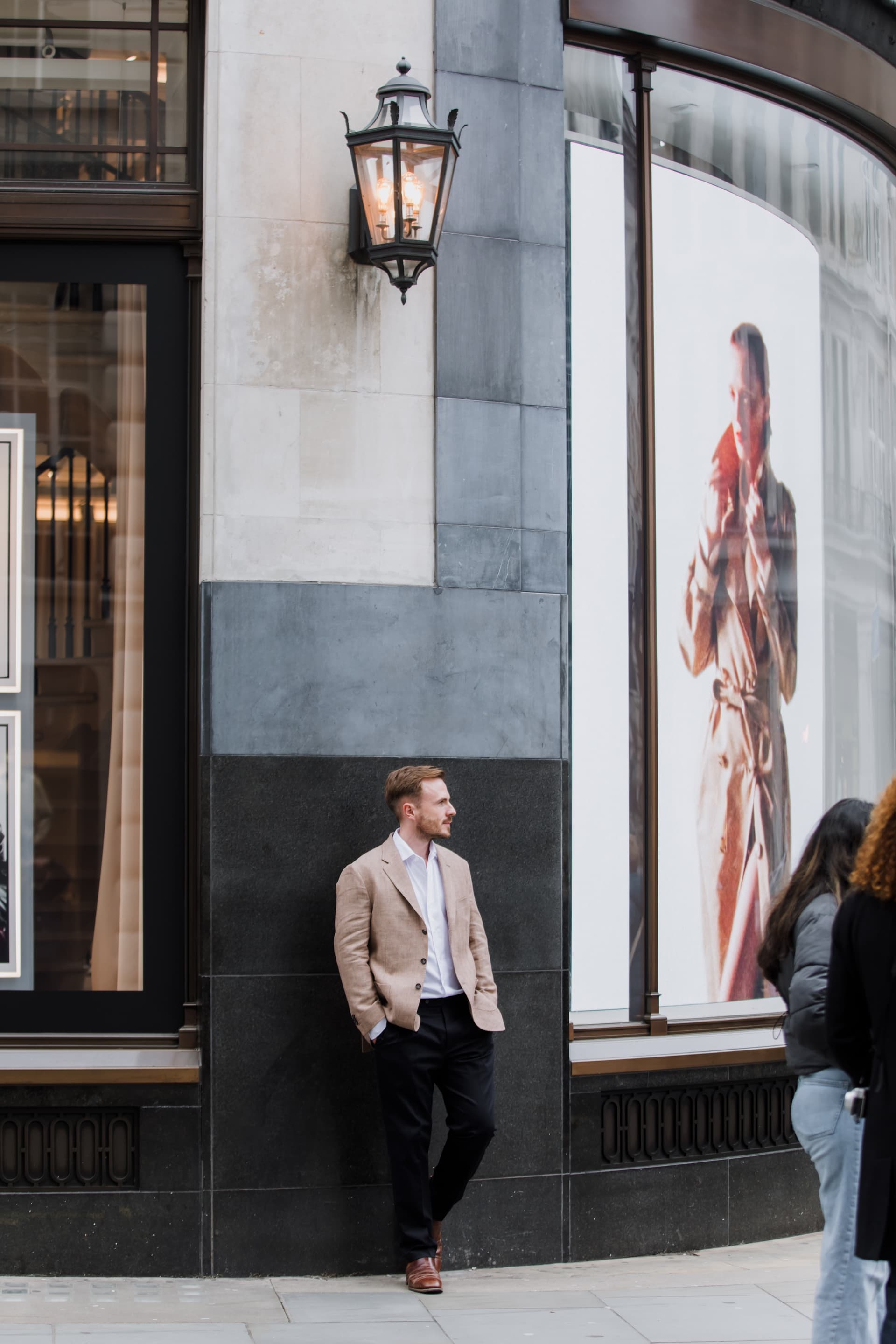 Dr Ben Davis in a tan linen blazer leaning against the corner of a shopfront beneath a lit Victorian lantern, looking away from the camera, on a London street