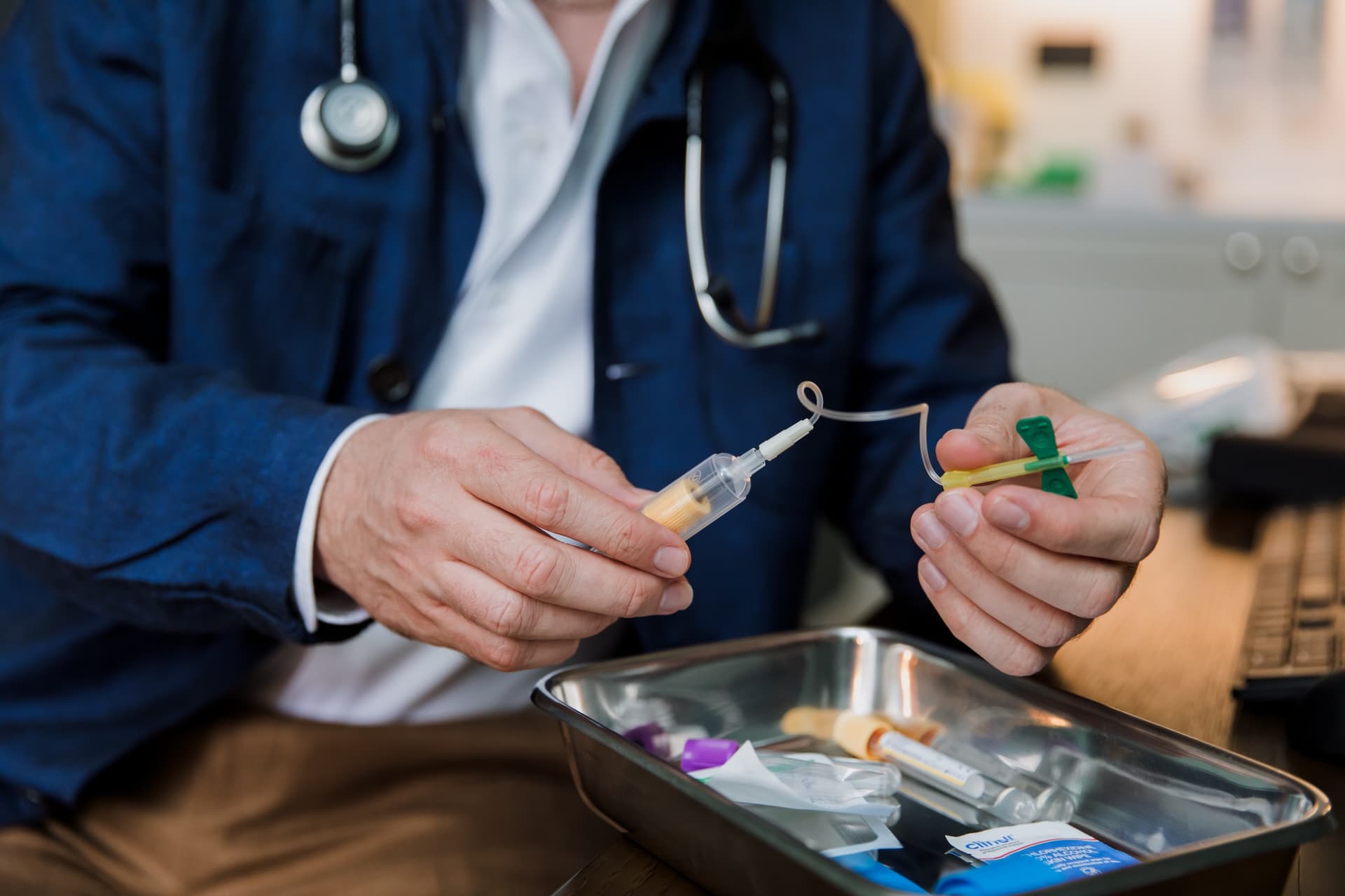Both hands holding a butterfly needle and blood collection set over a steel kidney dish, at the consulting room desk
