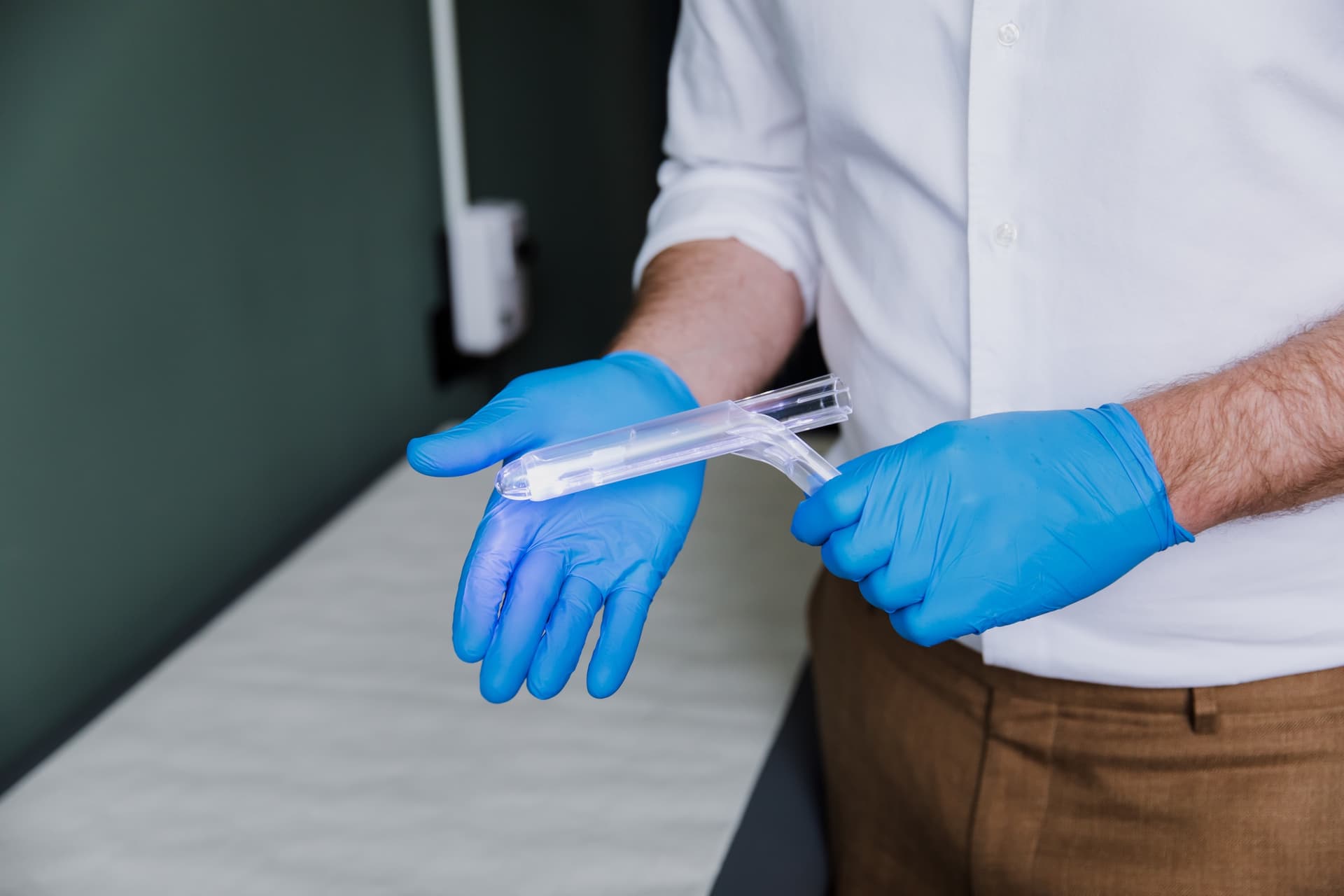 Gloved hands holding a proctoscope with its light on, beside the examination couch