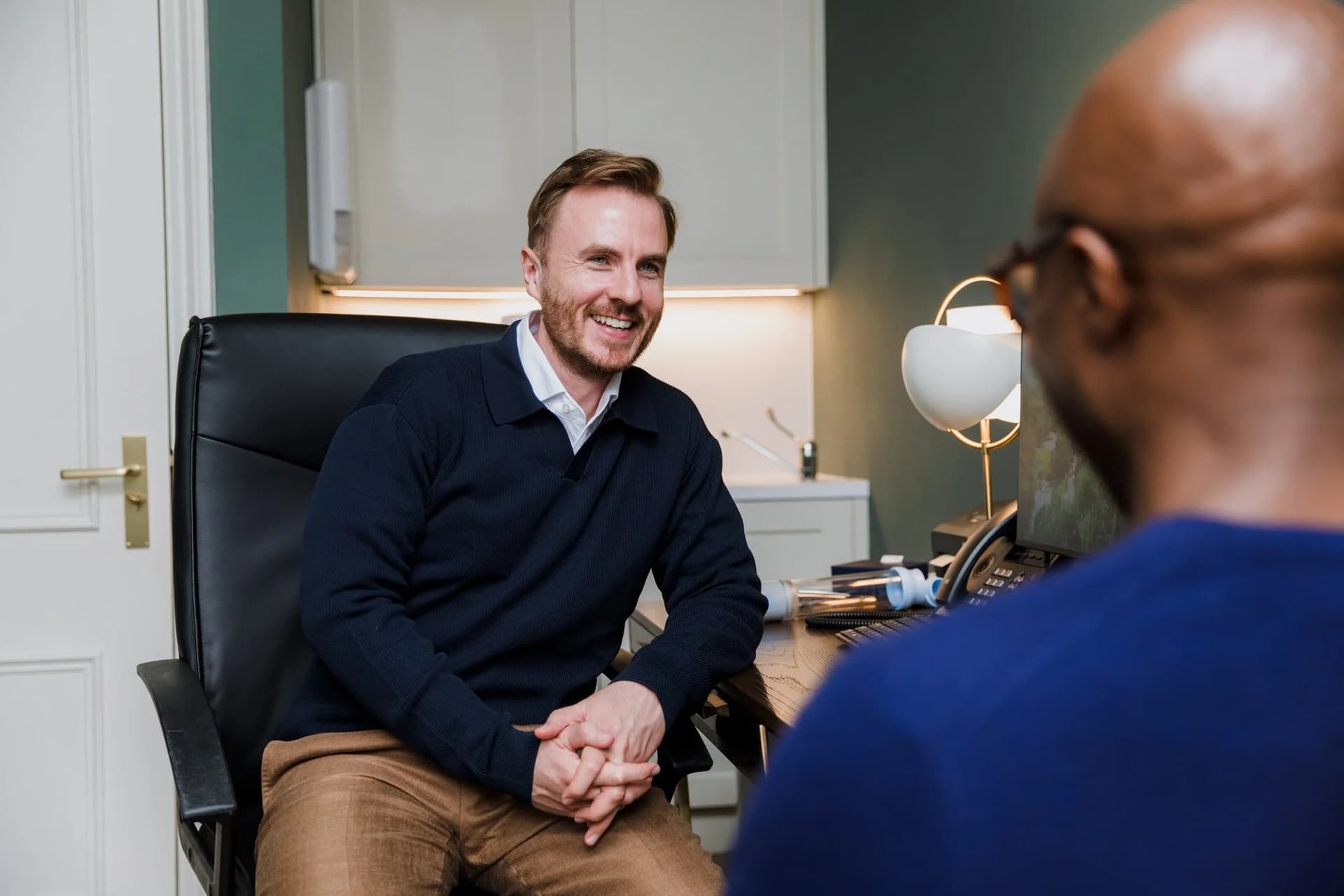 Dr Ben Davis listening, hands clasped, to a patient sitting opposite with his back to the camera