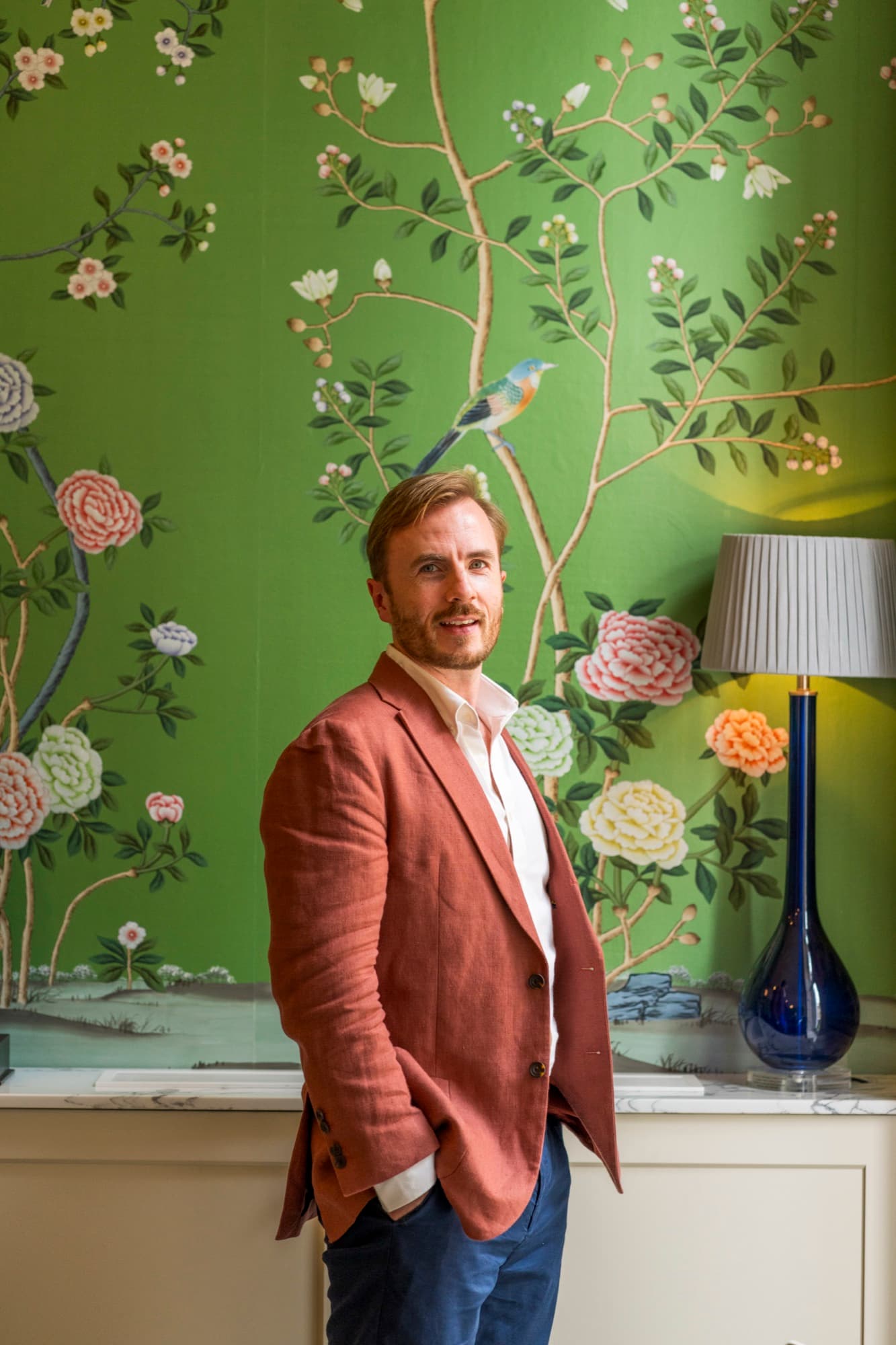 Dr Ben Davis standing in a rust linen jacket in front of hand-painted wallpaper, a green ground with blossoming branches, peonies and a small bird, beside a navy glass lamp