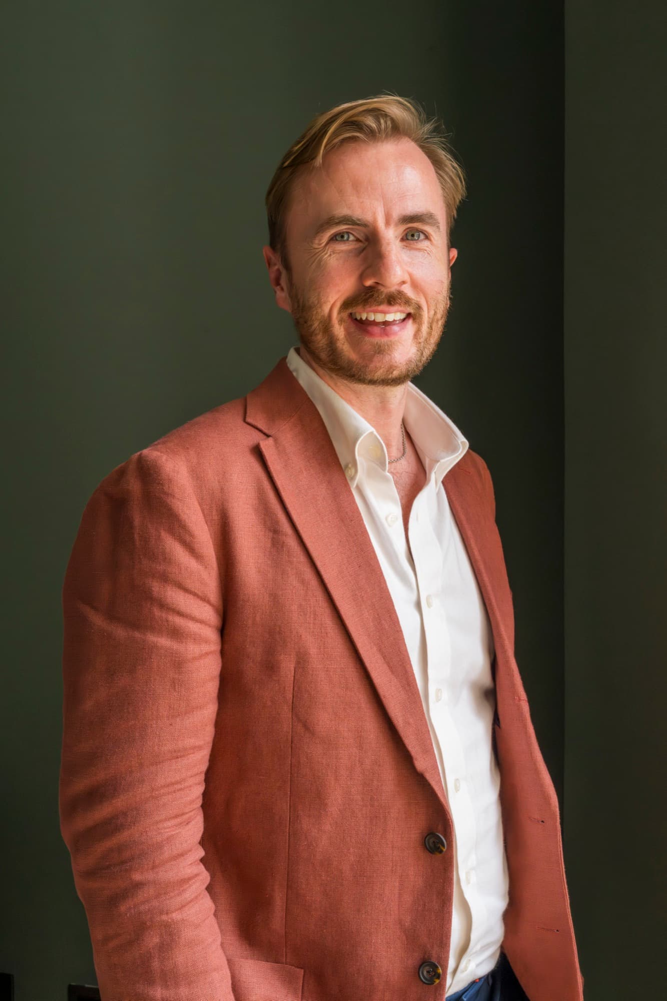 Dr Ben Davis smiling, in a rust linen jacket over an open cream shirt, against a dark olive wall lit from one side