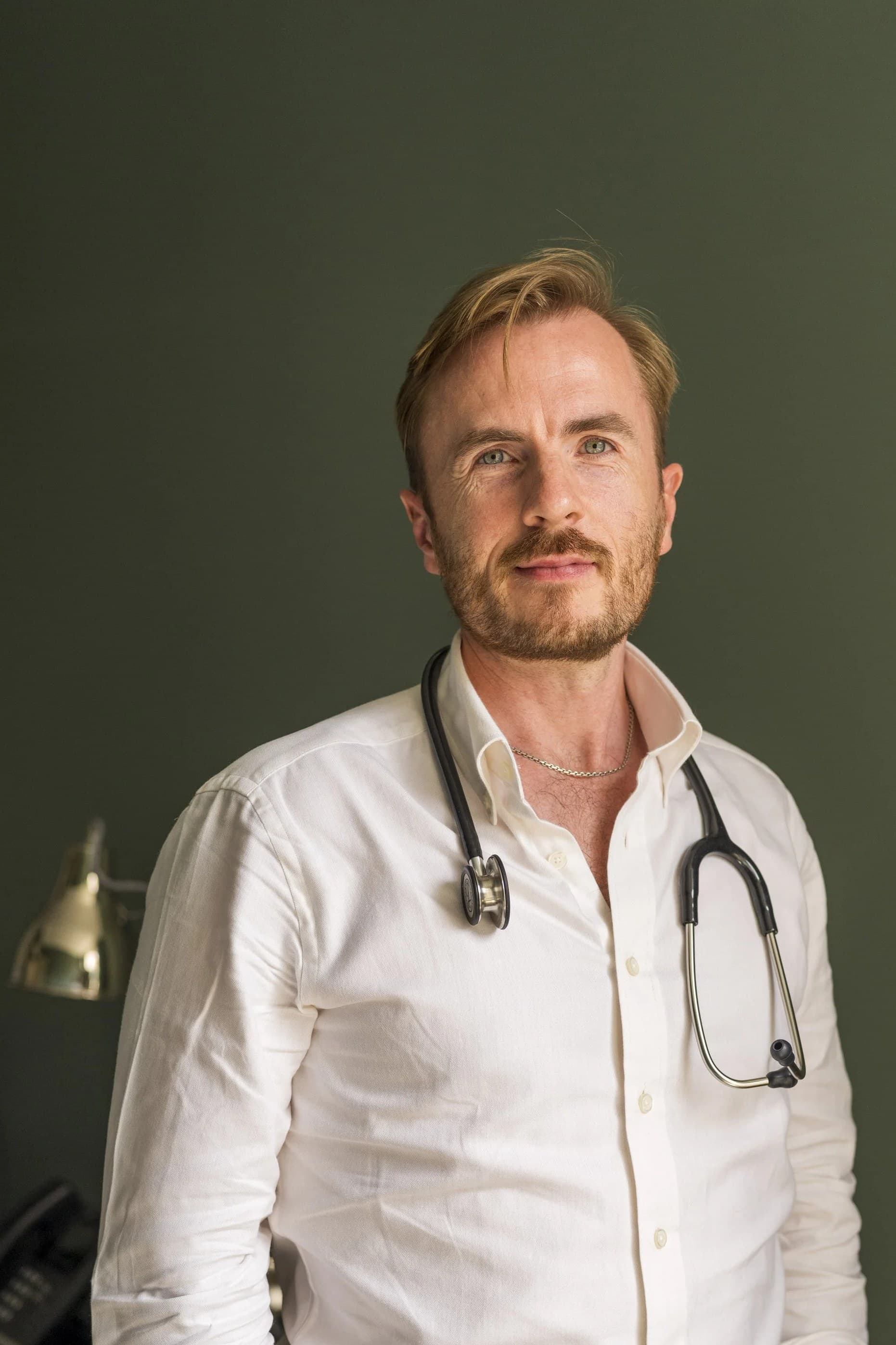 Dr Ben Davis standing in his consulting room in a white shirt with a stethoscope round his neck, against an olive green wall
