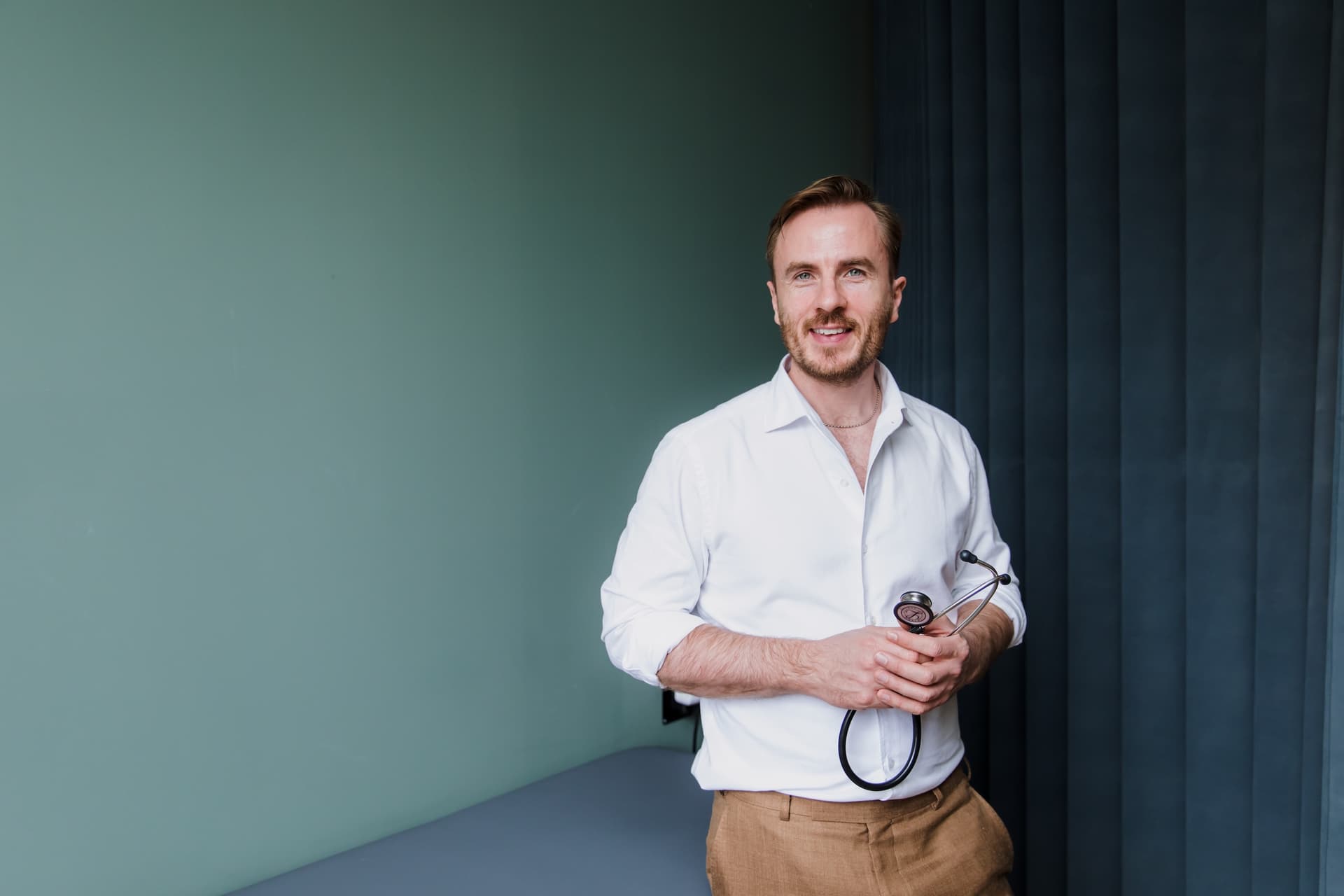 Dr Ben Davis standing against a soft green wall in a white shirt with the sleeves rolled, holding a stethoscope in both hands, beside the examination couch