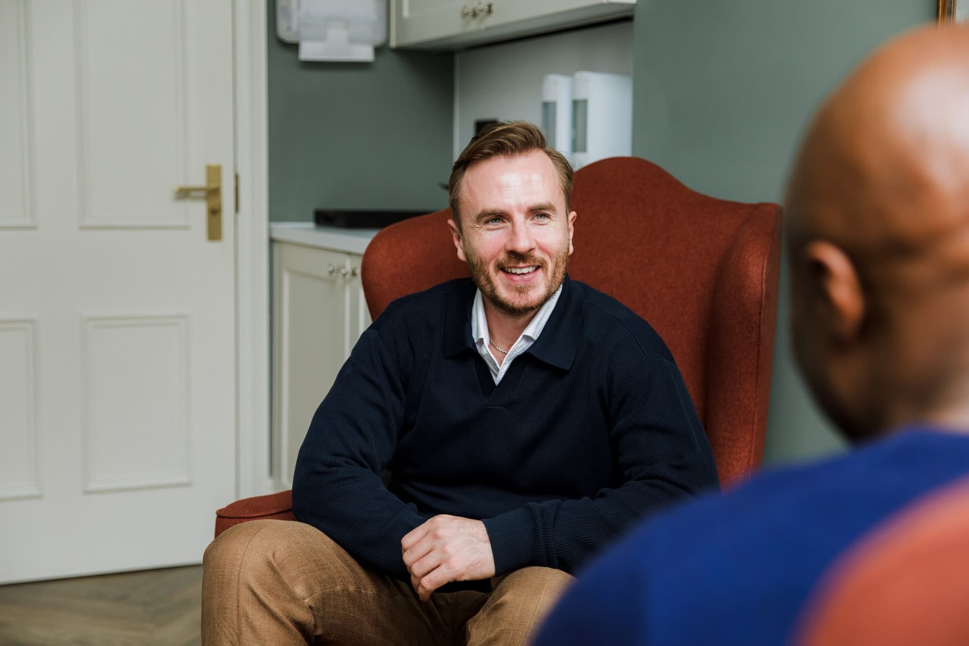 Dr Ben Davis sitting back in a red armchair, laughing, with a patient in the foreground seen from behind and out of focus