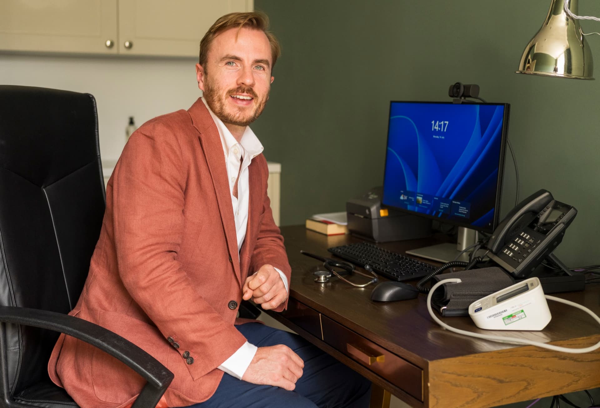 Dr Ben Davis turning from his desk to face the camera, with a monitor, telephone, stethoscope and blood-pressure monitor on the desk beside him