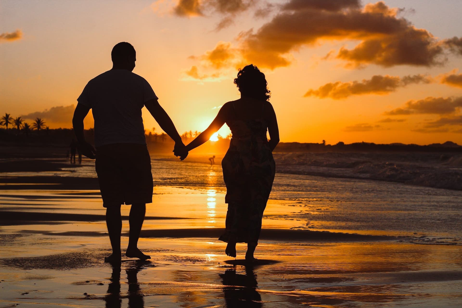 Two people walking hand in hand along a beach at sunset, seen in silhouette.