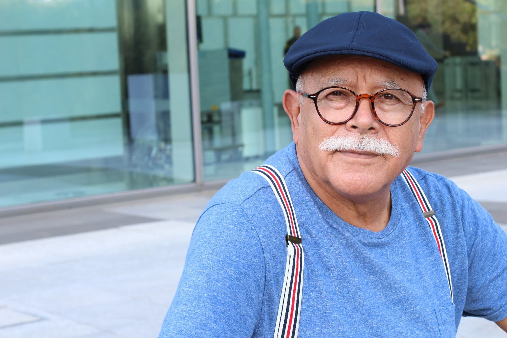 An older man in a flat cap, glasses and braces sitting outside, looking directly at the camera.