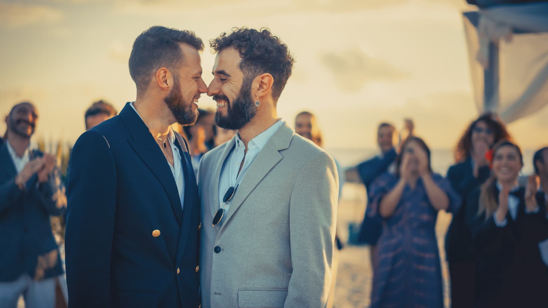 Two grooms in suits touching noses at an outdoor wedding while guests applaud behind them.