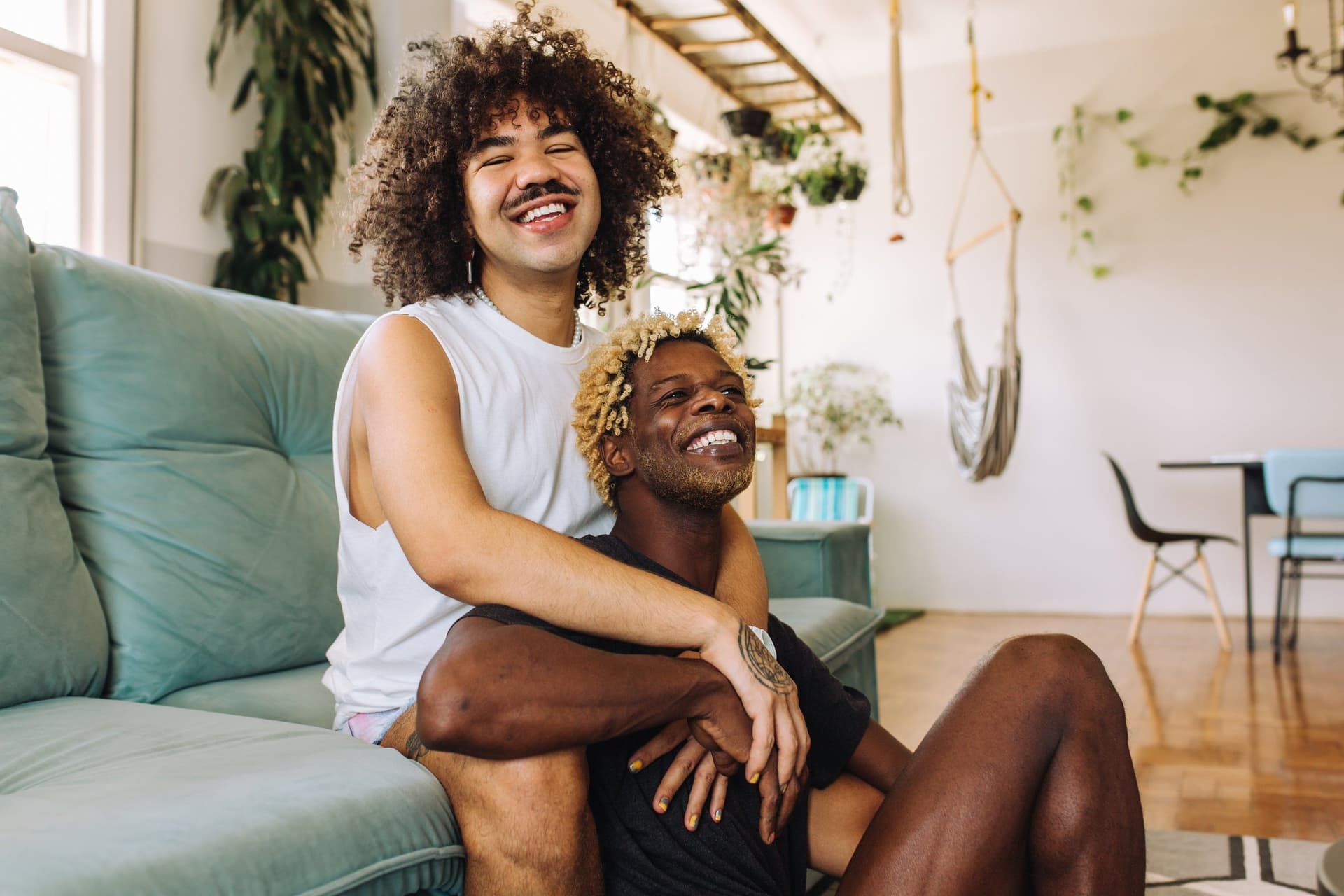 Two young men laughing together on a sofa at home, one with his arms around the other.