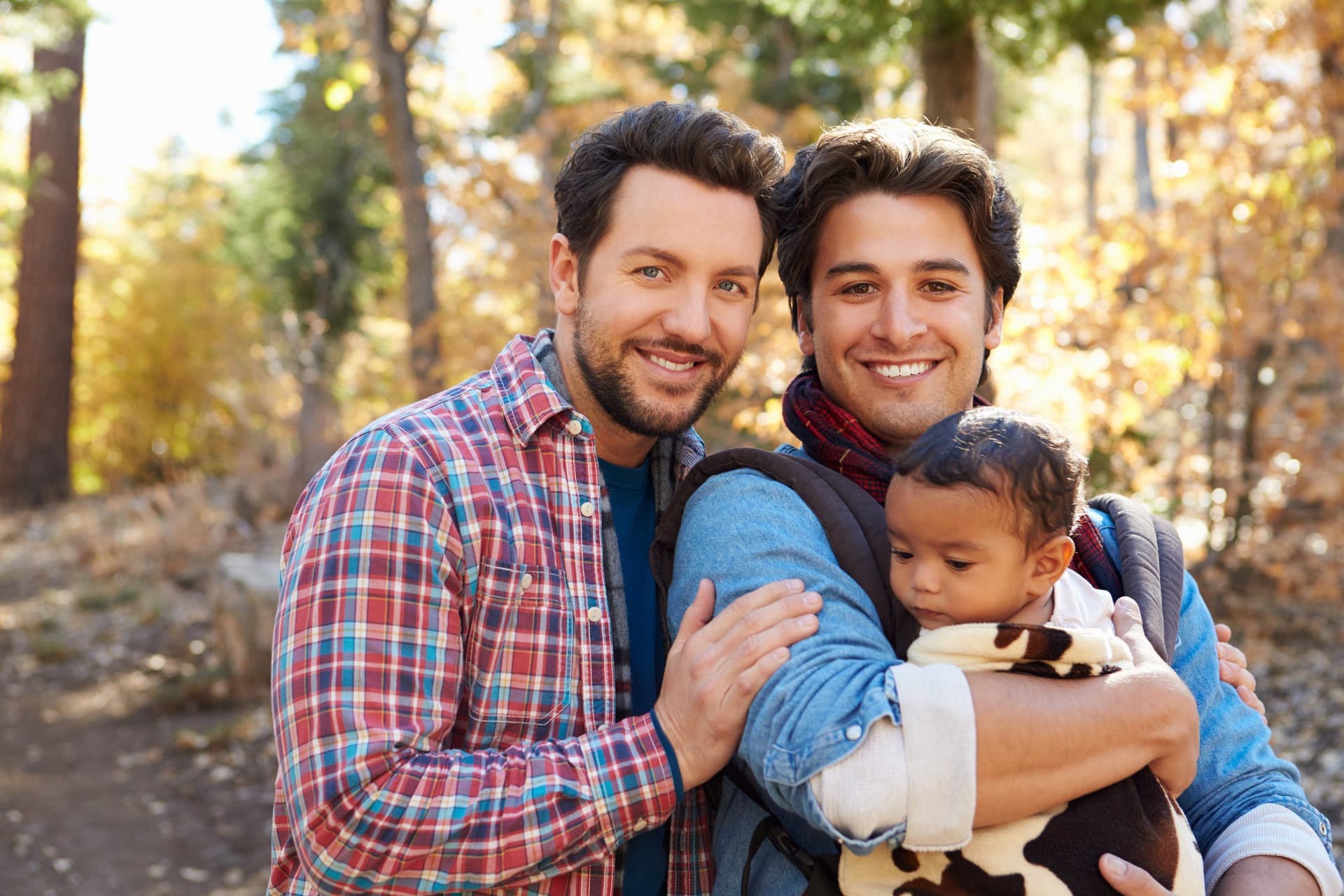 Two men standing close together in autumn woodland, one carrying a baby in a sling.